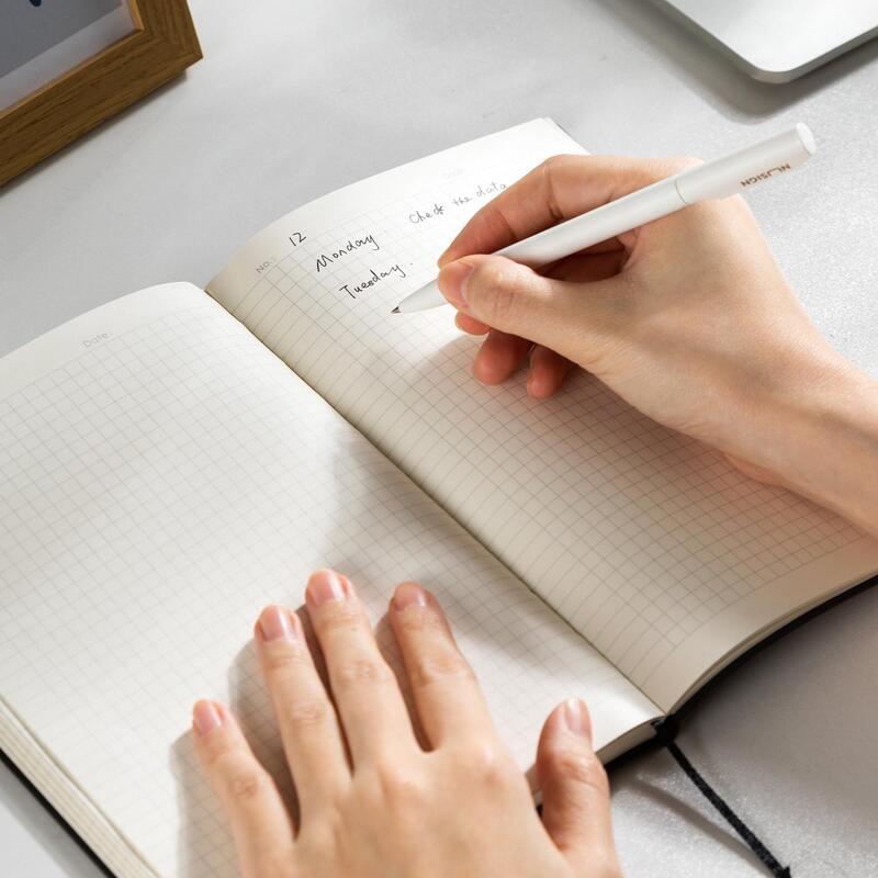 Hands writing in an open grid-paper notebook on a light gray desk next to a laptop